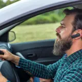 Mężczyzna z brodą i słuchawką Bluetooth w uchu prowadzi samochód. Dowiedz się, jak sparować telefon z samochodem, by bezpiecznie rozmawiać.
