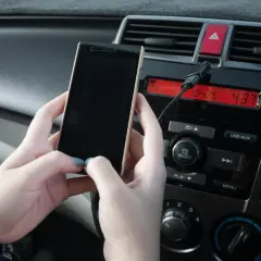 Ręce trzymają telefon, który jest podłączony kablem do radia samochodowego. Prosty sposób, jak połączyć telefon z samochodem.