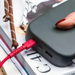 Dłoń z czerwonym lakierem podłącza czerwony kabel do powerbanku, zapewniając szybkie ładowanie.