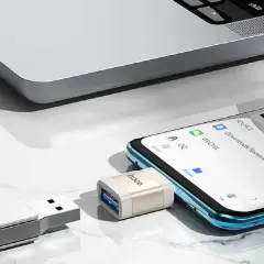 Adapter OTG podłączony do telefonu, umożliwiający transfer danych. Obok pendrive i fragment laptopa.