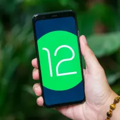 Dłoń trzyma smartfon z logo Android 12 na ekranie, na tle zieleni.