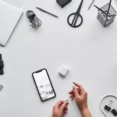 Ręce trzymające słuchawki AirPods obok telefonu z krajobrazem, sugerując, czy airpods działają z androidem. Na biurku laptop, kawa, nożyczki.