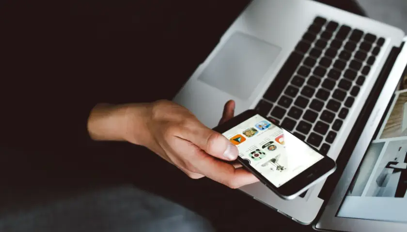 Dłoń trzyma iPhone'a z widocznymi ikonami aplikacji, obok otwartego laptopa. To idealny moment na **backup iPhone'a**.