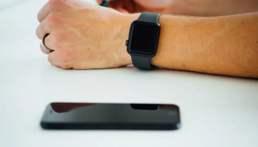 Czarny smartwatch na nadgarstku i leżący obok smartfon. Dowiedz się, jak włączyć bluetooth w zegarku, by połączyć go z telefonem.