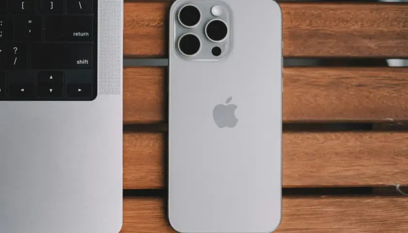 Srebrny iPhone 13 Pro z systemem iOS 26.4 obok klawiatury MacBook'a na drewnianym stole.
