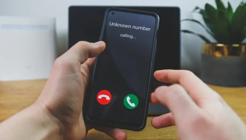Dłoń trzyma smartfon z ekranem pokazującym "Unknown number calling..." oraz przyciski odbierania i odrzucania połączenia.