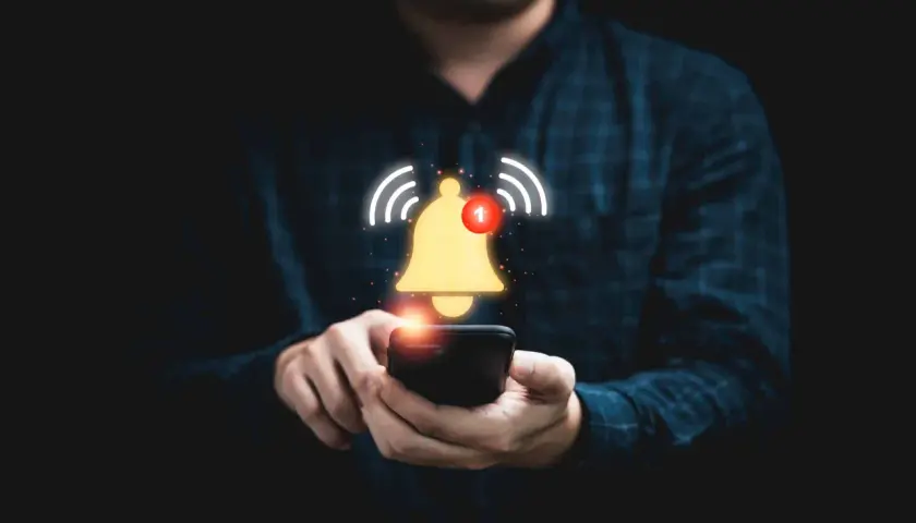 Dłoń trzymająca smartfon, nad którym unosi się złoty dzwonek z powiadomieniem.