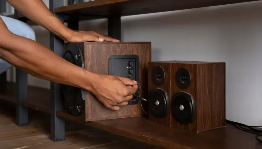 Ręka podłącza kabel do głośnika, pokazując, jak podłączyć subwoofer. Drewniane obudowy głośników stoją na półce.