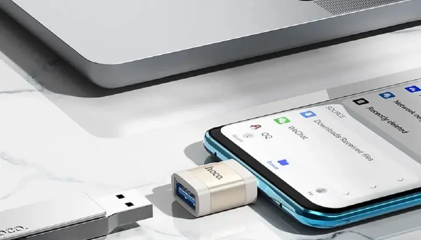 Adapter OTG podłączony do telefonu, umożliwiający transfer danych. Obok pendrive i fragment laptopa.