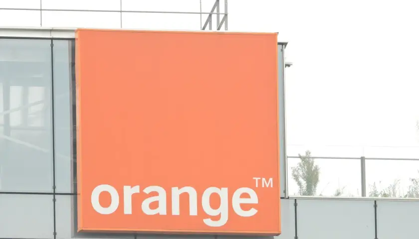 Pomarańczowy znak z napisem "orange" na nowoczesnym budynku. To logo firmy telekomunikacyjnej, która oferuje usługi multiroom.