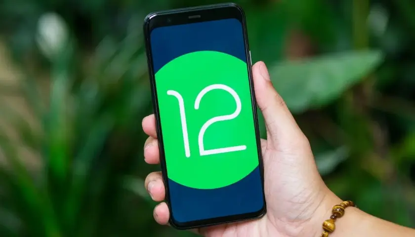 Dłoń trzyma smartfon z logo Android 12 na ekranie, na tle zieleni.