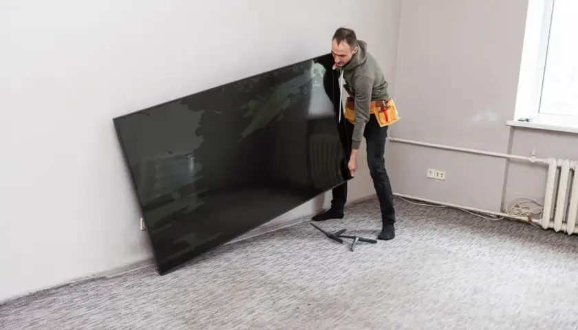 Mężczyzna z pasem narzędziowym przenosi duży, czarny ekran. To telewizor o wymiarach 55 cali, gotowy do montażu.