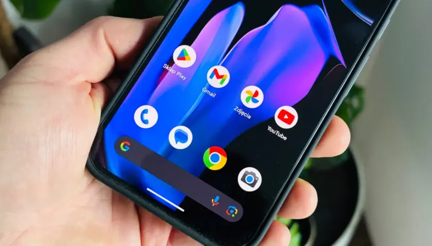 Dłoń trzyma smartfon z kolorową tapetą i ikonami aplikacji, w tym Sklep Play, Gmail, Zdjęcia i YouTube. Widoczny pasek wyszukiwania Google i ikona aparatu. To może być zapowiedź funkcji w nadchodzącym android 15.