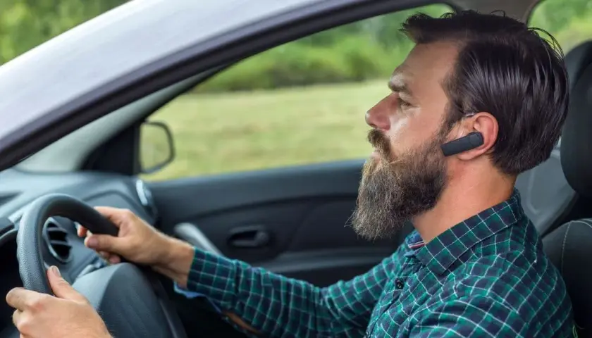 Mężczyzna z brodą i słuchawką Bluetooth w uchu prowadzi samochód. Dowiedz się, jak sparować telefon z samochodem, by bezpiecznie rozmawiać.