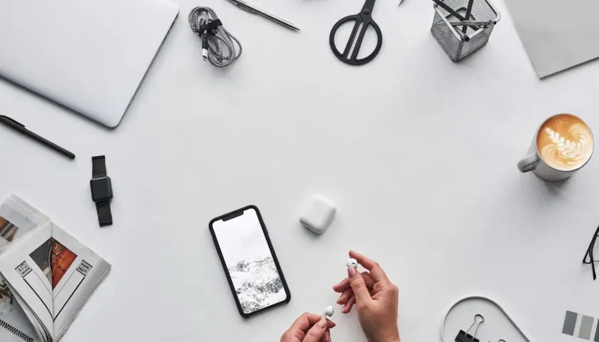 Ręce trzymające słuchawki AirPods obok telefonu z krajobrazem, sugerując, czy airpods działają z androidem. Na biurku laptop, kawa, nożyczki.