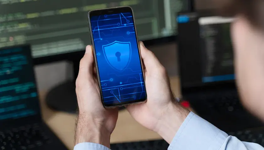 Ręce trzymające smartfon z ikoną tarczy i klucza, symbolizującą jak odblokować telefon. W tle monitory z kodem.