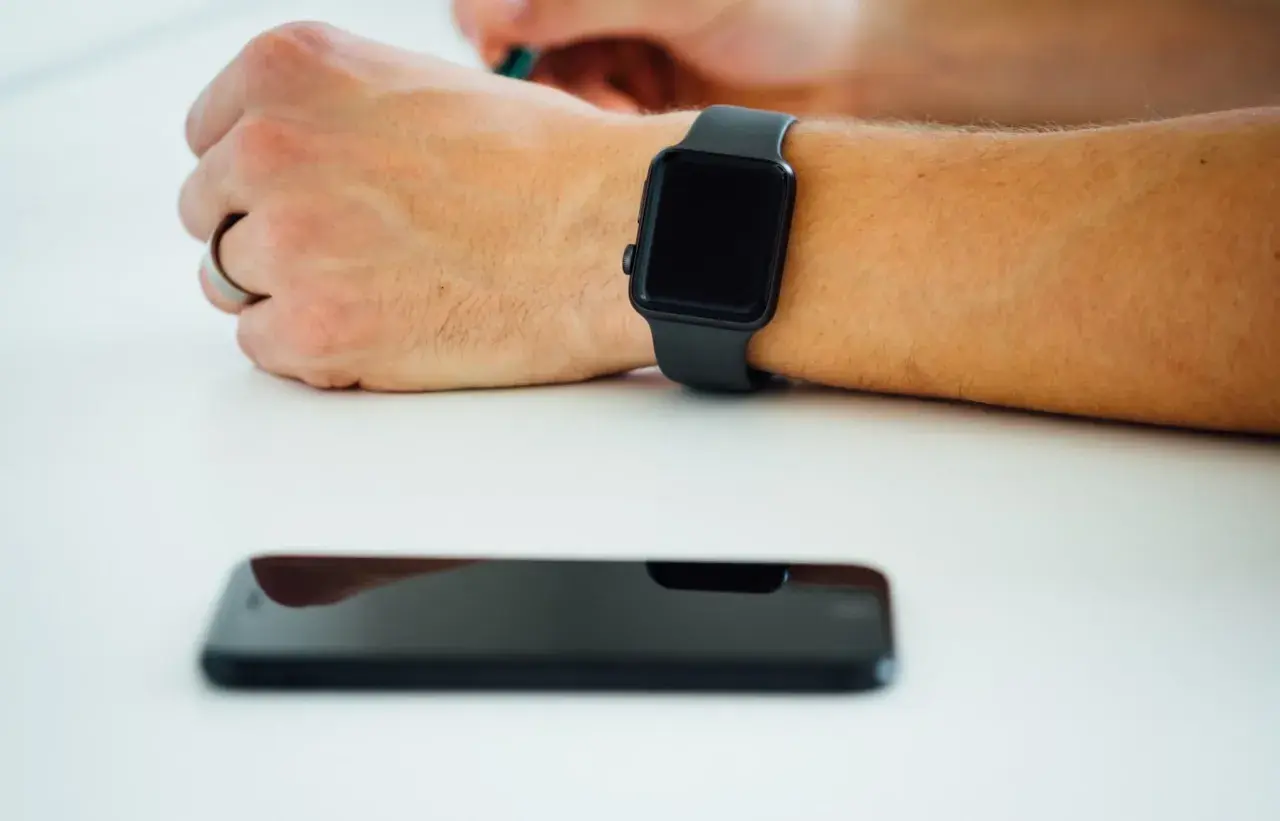 Czarny smartwatch na nadgarstku i leżący obok smartfon. Dowiedz się, jak włączyć bluetooth w zegarku, by połączyć go z telefonem.