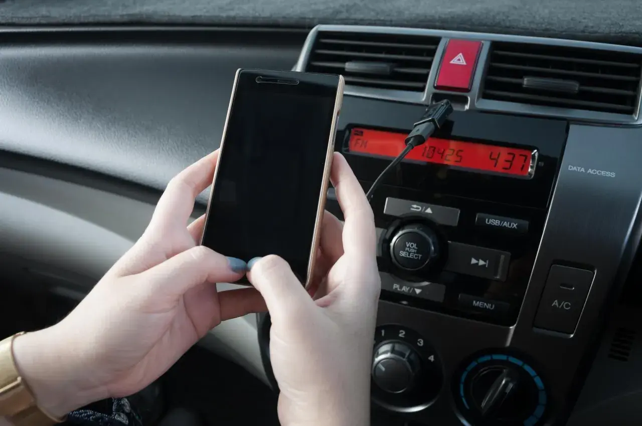Ręce trzymają telefon, który jest podłączony kablem do radia samochodowego. Prosty sposób, jak połączyć telefon z samochodem.