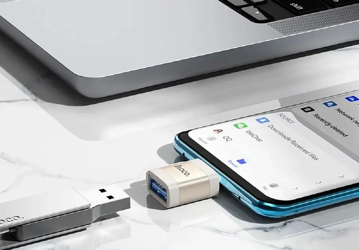 Adapter OTG podłączony do telefonu, umożliwiający transfer danych. Obok pendrive i fragment laptopa.