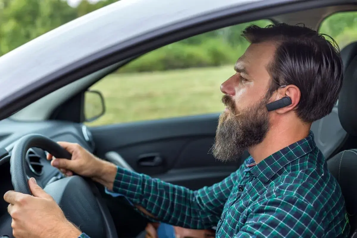 Mężczyzna z brodą i słuchawką Bluetooth w uchu prowadzi samochód. Dowiedz się, jak sparować telefon z samochodem, by bezpiecznie rozmawiać.