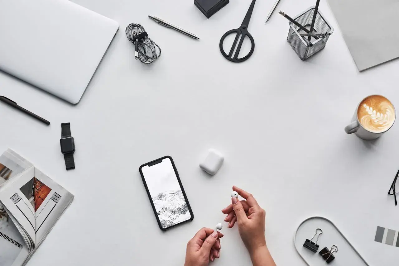 Ręce trzymające słuchawki AirPods obok telefonu z krajobrazem, sugerując, czy airpods działają z androidem. Na biurku laptop, kawa, nożyczki.