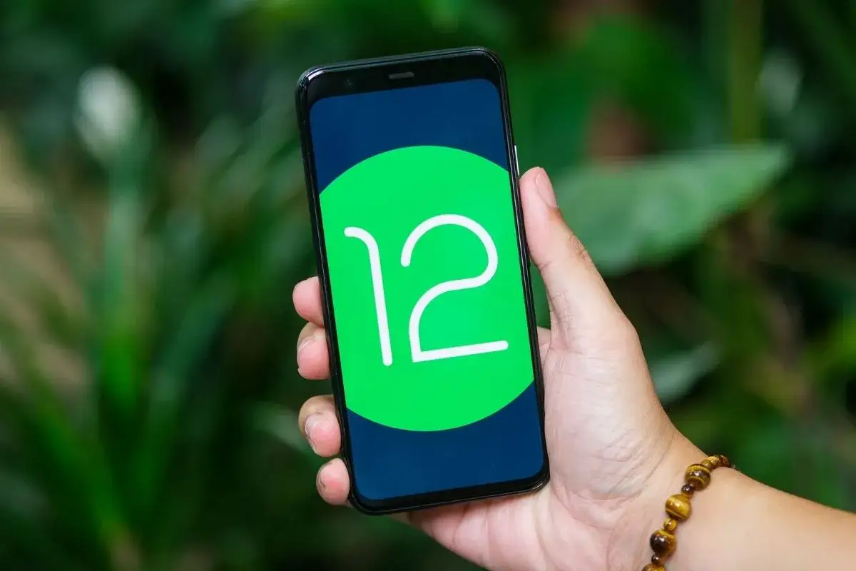 Dłoń trzyma smartfon z logo android 12 na ekranie. Tło to rozmyte zielone liście.